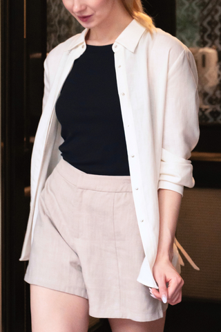 A woman wearing the Olive Basics Marie Everyday Shirt in TENCEL™ Linen (white) over a black top and beige shorts, walking indoors.