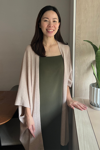 A woman with long dark hair stands indoors beside a plant, smiling at the camera while wearing a green dress and the Olive Basics Deborah Nursing Kimono Cardigan in light beige.