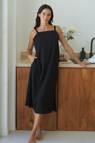 A woman stands barefoot in a kitchen, leaning against a wooden counter with flowers, wearing the Olive Basics Hannah Reversible Midi Dress.
