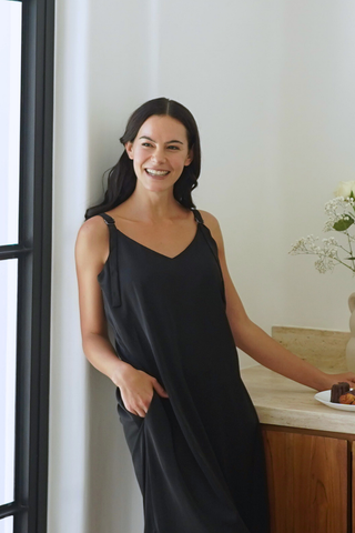 A woman wears the Olive Basics Hannah Reversible Midi Dress indoors, smiling by a wooden counter with pastries and a vase of flowers nearby.