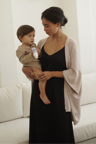 A woman in the Olive Basics Hannah Reversible Midi Dress and light cardigan holds a young child in beige, who holds a small cylindrical object, standing together in a neutral-toned room.
