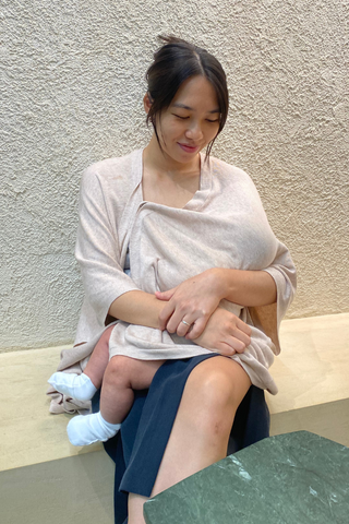A woman breastfeeds an infant on a bench, partially covered by the Olive Basics Deborah Nursing Kimono Cardigan, with a textured wall in the background.