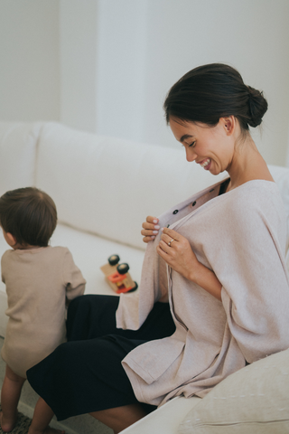 A woman sits on a couch, smiling as she adjusts her Olive Basics Deborah Nursing Kimono Cardigan. A small child with toy cars sits beside her, facing away.
