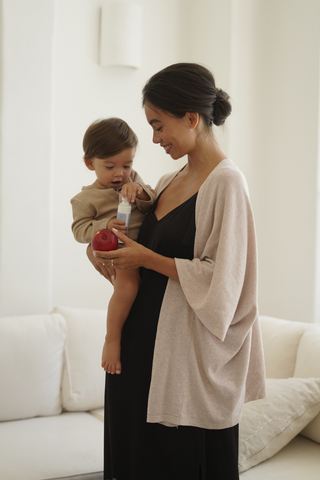 A woman wearing the Olive Basics Deborah Nursing Kimono Cardigan holds a young child in a bright living room, offering a bottle while the child holds a red apple.