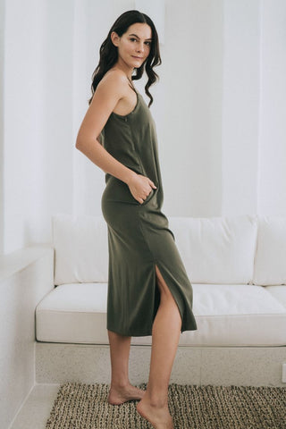 A woman stands barefoot on a rug in front of a white couch, wearing the Olive Basics Hannah Reversible Midi Dress—an olive green A-line style with a reversible neckline and side slit.