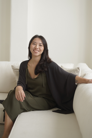 A woman with long dark hair sits on a light couch, wearing an olive green dress and the Deborah Nursing Kimono Cardigan by Olive Basics, smiling at the camera.