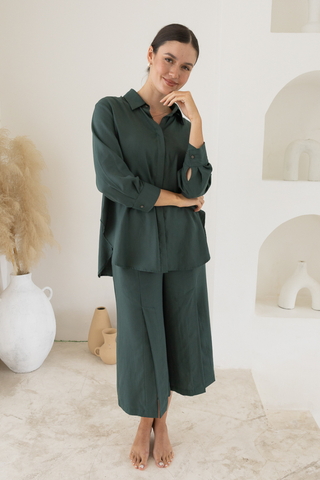 A woman stands indoors, barefoot, in Olive Basics’ Delilah All-Occasion TENCEL™ Shirt and matching wide-leg pants; her neutral expression and hand to face highlight the breastfeeding-friendly design for versatile wear.