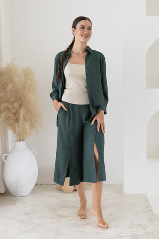 A woman with a braid wears Olive Basics Delilah All-Occasion TENCEL™ Shirt in dark green, styled with a matching skirt over a beige top, standing barefoot in a minimalist room with neutral decor.