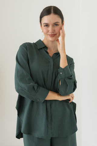 A person with dark hair in a low bun wears the loose-fitting Delilah All-Occasion TENCEL™ Shirt from Olive Basics, paired with matching pants, standing against a plain white background and touching their face with one hand.