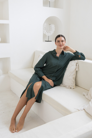 A woman in the Olive Basics Delilah All-Occasion TENCEL™ Shirt, long-sleeved and dark green, sits barefoot on a white cushioned bench in a minimalist, white-walled room.