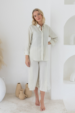 A woman stands barefoot indoors in a loose, cream-colored Delilah All-Occasion TENCEL™ Shirt and matching wide-leg pants from Olive Basics, with neutral pottery placed beside her.