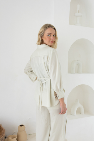 A woman with blonde hair stands indoors wearing the Olive Basics Delilah All-Occasion TENCEL™ Shirt, breastfeeding friendly and light-colored, paired with pants. Minimalist white decor and ceramics appear in the background.
