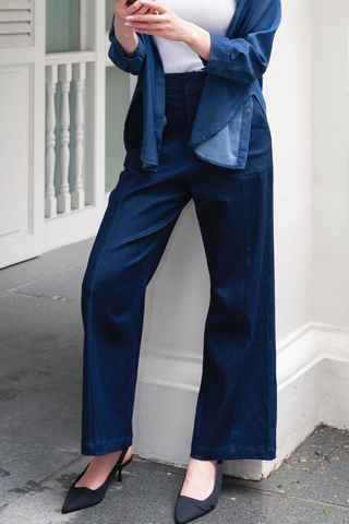 A person wearing a blue shirt, Olive Basics Jesse Wide-leg Pocket Pants in Denim TENCEL, and black heels stands outside holding a smartphone. Their head is not visible in the image.