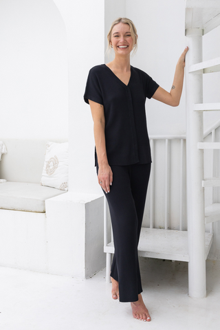 A woman stands barefoot indoors, smiling in the Olive Basics Eve Button-Down Knit Tee and matching black pants, with one hand resting on a white spiral staircase.
