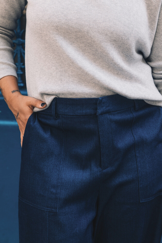 A person wears a light gray ribbed sweater and Olive Basics Jesse Wide-leg Pocket Pants in Denim TENCEL, with one hand in a pocket, standing before a blue background.