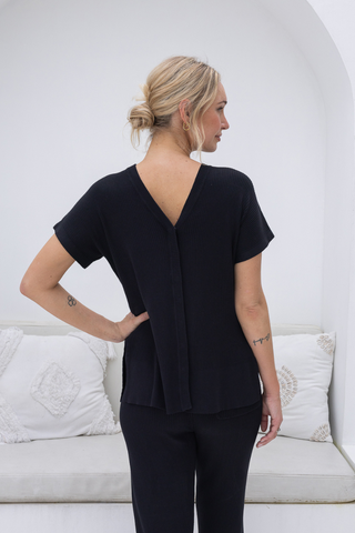 A person with blonde hair in a low bun, seen from behind indoors, wears the Olive Basics Eve Button-Down Knit Tee in black with matching pants.