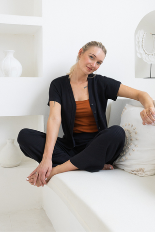 A woman sits cross-legged on a white cushioned bench, wearing Olive Basics Eve Button-Down Knit Tee in black over a brown top, which offers breastfeeding access. White decorative vases and jewelry are displayed in the background.