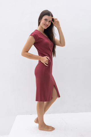 A woman stands barefoot on a white platform, wearing Olive Basics' Ruth Reversible T-shirt Dress—short-sleeved, knee-length maroon with a side slit—posing against a plain white background.