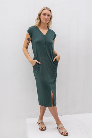 Woman in the Olive Basics Ruth Reversible T-shirt Dress, a dark green, button-front maternity midi with short sleeves and pockets, stands on a white platform against a white background.