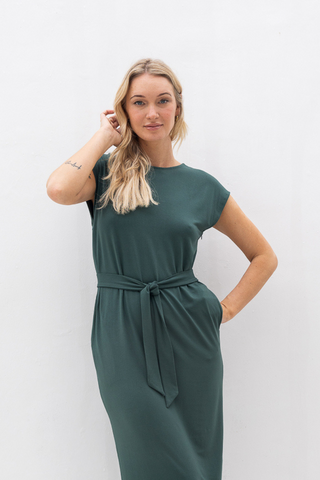 Woman wearing the Olive Basics Ruth Reversible T-shirt Dress, a dark green maternity style with a belt, standing against a white background with one hand in her pocket and the other touching her hair.