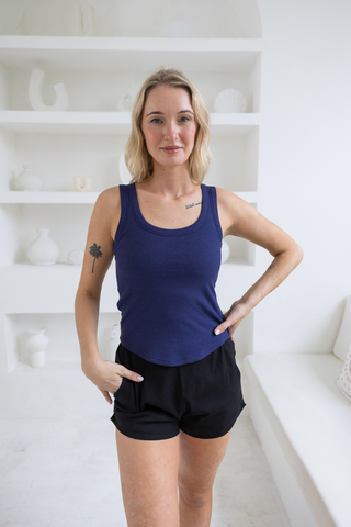 A blonde woman stands indoors, one hand on her hip and the other in her pocket, wearing the Olive Basics Anna Reversible Hi-Low Tank in blue with black shorts. White shelves with decor are visible in the background.