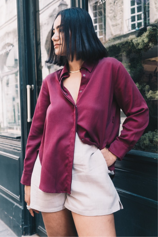 A woman with shoulder-length dark hair wears the Olive Basics Marie Everyday Shirt in TENCEL™ (burgundy) and light-colored shorts, standing outside in front of a dark storefront.