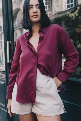 A woman stands outdoors, wearing Olive Basics Joanna Expandable Pocket Shorts in TENCEL™ Linen—a high-waisted style with adjustable waistband—paired with a burgundy button-up shirt, one hand in her pocket and a neutral expression.