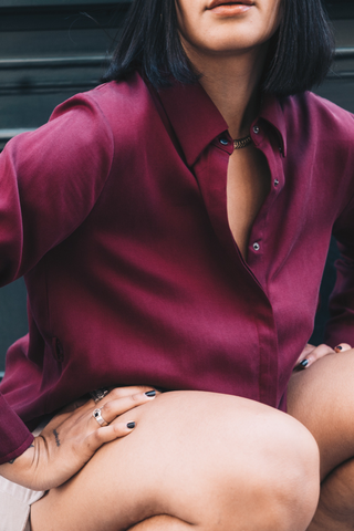 A person with shoulder-length dark hair wears the Marie Everyday Shirt in TENCEL™ by Olive Basics, paired with beige shorts and jewelry, sitting with hands on knees.
