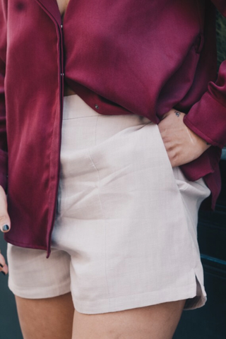A person wears Olive Basics Joanna Expandable Pocket Shorts in TENCEL™ Linen, high-waisted with hands in pockets, paired with a burgundy satin shirt, shown from the waist down.