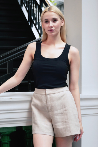 A woman with long blonde hair stands indoors by a staircase, wearing a black sleeveless top and Olive Basics Joanna Expandable Pocket Shorts in TENCEL™ Linen with an adjustable waistband, looking at the camera.