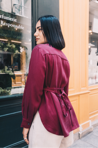 A woman with straight black hair stands outside, facing away, wearing the Marie Everyday Shirt in TENCEL™ by Olive Basics in maroon with long sleeves and a belt, paired with light pants.