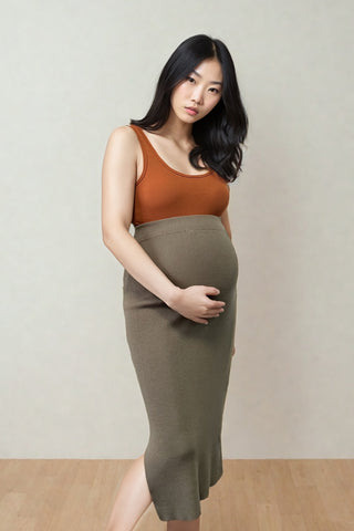 A pregnant woman with long dark hair stands indoors, gently holding her belly, wearing an orange tank top and the Olive Basics Leah Super Stretch Slit Skirt.