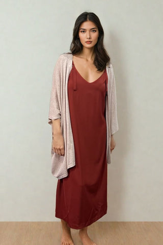 A woman stands barefoot on a wooden floor in the Olive Basics Hannah Reversible Midi Dress, a long, sleeveless maroon A-line style, paired with a light pink open-front cardigan against a plain light background.