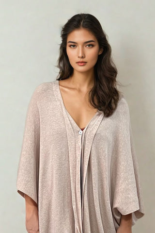 A woman with long, wavy dark hair stands against a plain light background, wearing the Olive Basics Deborah Nursing Kimono Cardigan in a loose-fitting, light beige style with a deep neckline and buttons.