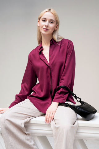 A woman with light hair sits on a white surface, wearing the Marie Everyday Shirt in TENCEL™ by Olive Basics in burgundy, beige pants, and holding a black handbag.