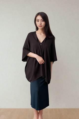 A woman stands indoors against a plain light wall, wearing the Olive Basics Deborah Nursing Kimono Cardigan in black and a dark skirt, with her hands resting in front.