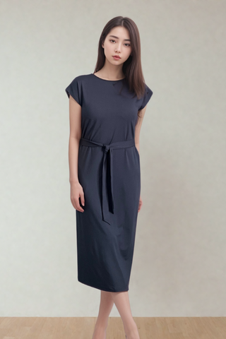 A woman stands indoors in the Olive Basics Ruth Reversible T-shirt Dress, a dark, short-sleeved breastfeeding dress with a belt, paired with neutral heels against a plain light background.