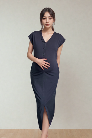 A woman stands indoors wearing the Olive Basics Ruth Reversible T-shirt Dress, a dark, short-sleeved maternity outfit, with one hand on her abdomen and the other behind her back.