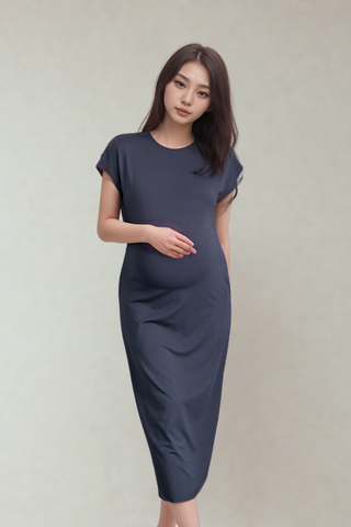 A woman in the Olive Basics Ruth Reversible T-shirt Dress, a dark blue short-sleeved maternity style, stands against a plain background with one hand on her baby bump.
