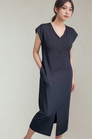 A woman stands against a plain background wearing the Olive Basics Ruth Reversible T-shirt Dress in grey blue—a midi maternity style with short sleeves, button front, V-neck, front slit, and pockets.