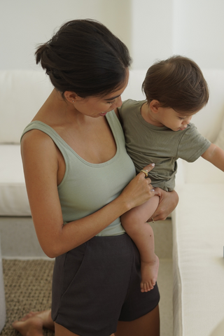 A woman in the Olive Basics Esther Easy Latch Tank, featuring a reversible neckline and built-in bra padding, holds a young child on her hip as they both look down at an object on a white table.