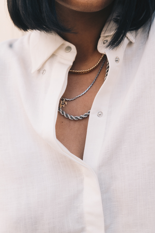 A person wears the Olive Basics Marie Everyday Shirt in TENCEL™ Linen (white), styled with three layered gold and silver chain necklaces.