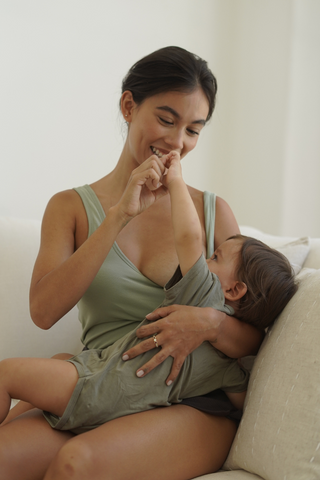 A woman wearing the Olive Basics Esther Easy Latch Tank sits on a couch, breastfeeding her young child and smiling as the child touches her face.