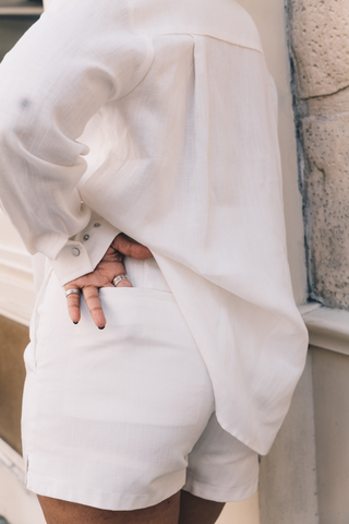 A person stands facing away with a hand on their lower back, wearing a loose white shirt and Olive Basics’ Joanna Expandable Pocket Shorts in TENCEL™ Linen with an adjustable waistband.