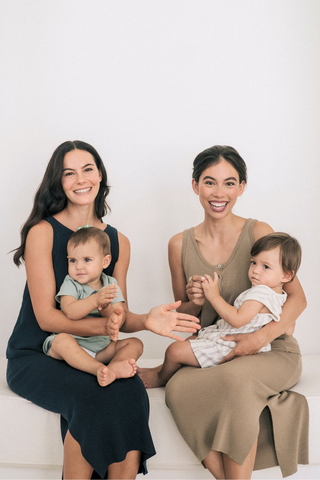 Two women sit side by side on a bench with toddlers on their laps, all smiling at the camera. Both wear the Leah Super Stretch Slit Skirt by Olive Basics, offering easy movement and style against the plain white background.