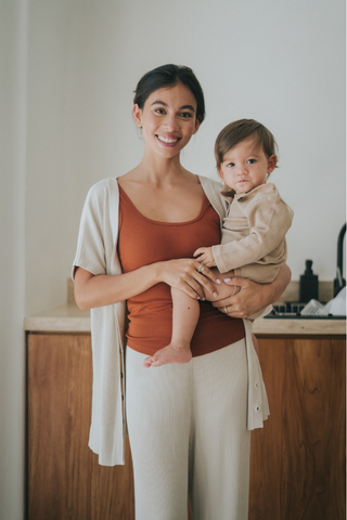 A woman in an Olive Basics Eve Button-Down Knit Tee—a rust-colored top with discreet breastfeeding access—holds a young child dressed in beige, standing in a modern kitchen. Ideal for busy moms.