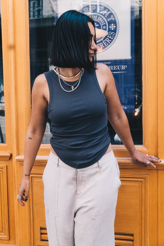 A woman with shoulder-length black hair wears the Olive Basics Anna Reversible Hi-Low Tank, light pants, and layered necklaces while standing in front of a wooden door.