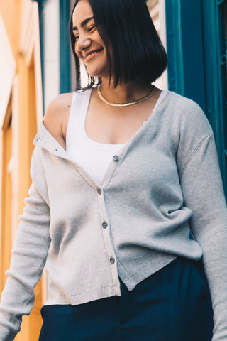 Wearing the Olive Basics Ellie Reversible Cardigan in TENCEL™ Knit over a white top and dark pants, a person with straight dark hair stands outdoors, smiling and looking down.