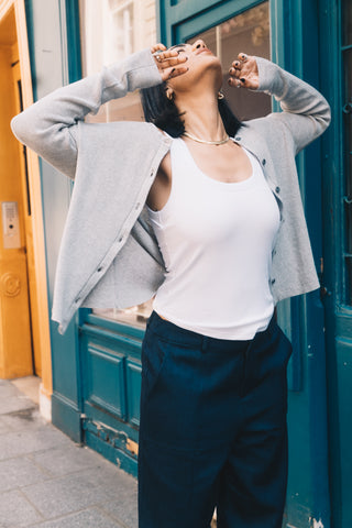 Wearing a white tank top, dark pants, and the Olive Basics Ellie Reversible Cardigan in TENCEL™ Knit, a person stands on a city sidewalk with their head tilted back and hands near their face.