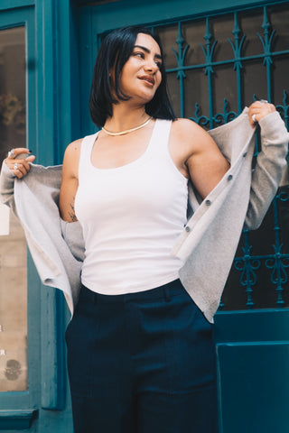 A woman wearing the Olive Basics Anna Reversible Hi-Low Tank and dark pants stands by a blue door, putting on a gray cardigan and smiling softly to the side.